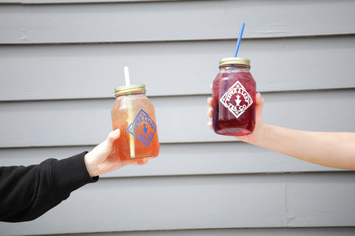 Two hands hold mason jar glasses filled with beverages against a light gray wooden wall.