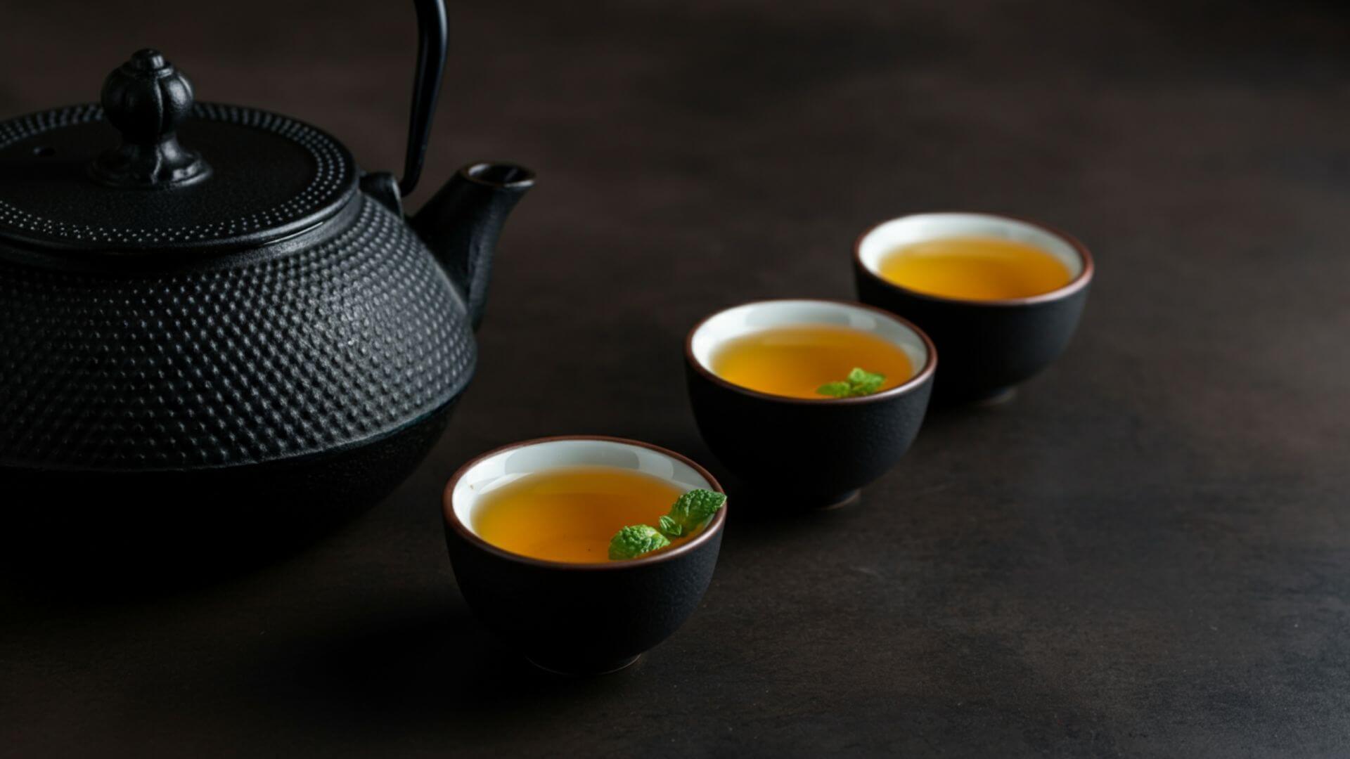 A black teapot sits on the left and in the center are three small tea cups filled with genmaicha tea