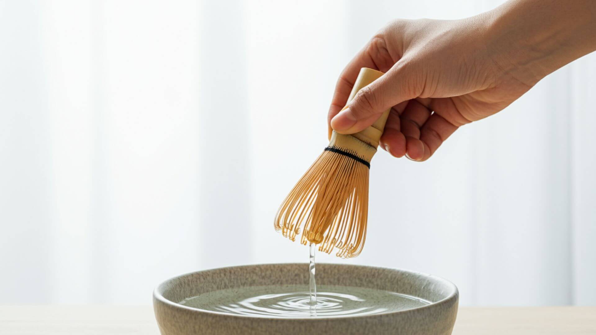 A hand delicately holds a bamboo whisk over a bowl with water