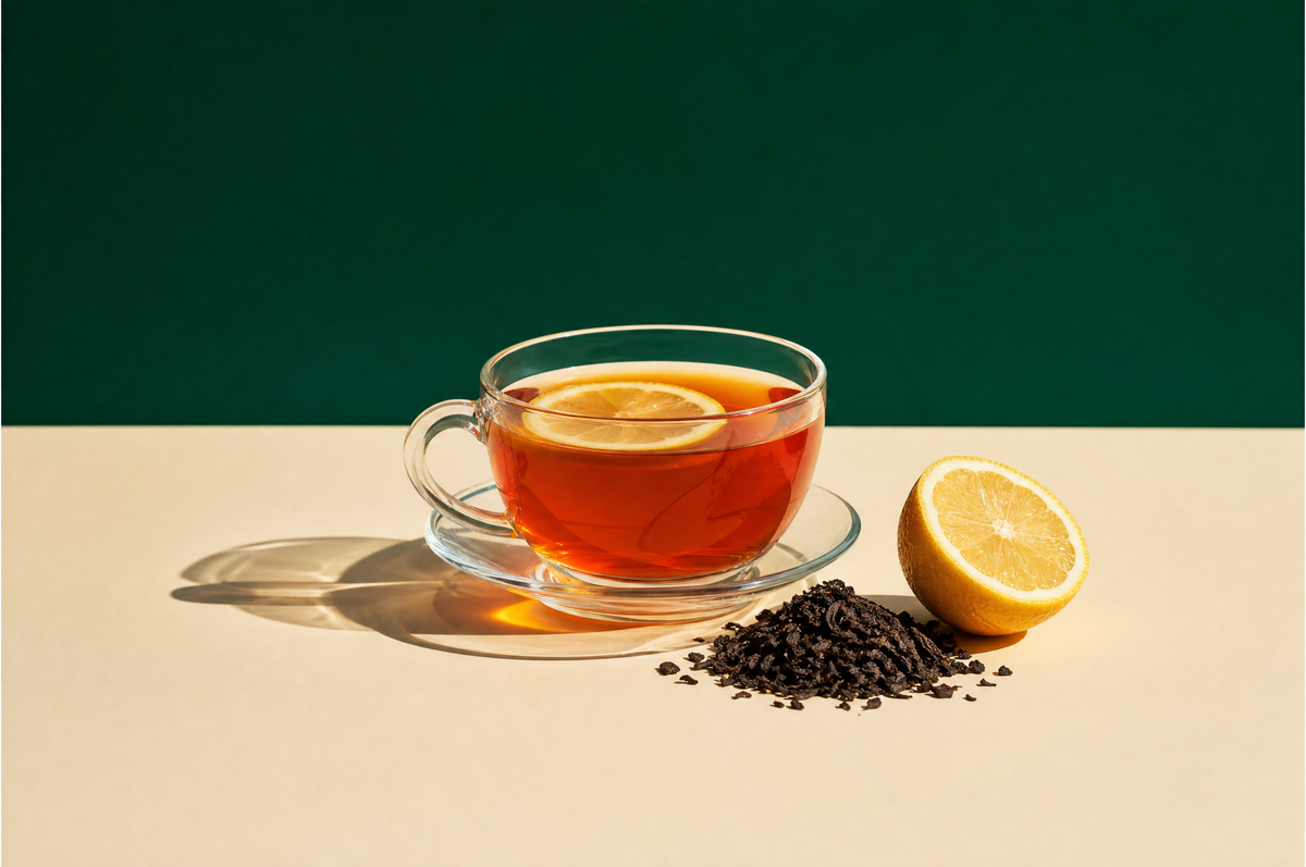 A clear glass teacup on a matching clear glass saucer filled with bright golden-red black tea with a thin lemon slice