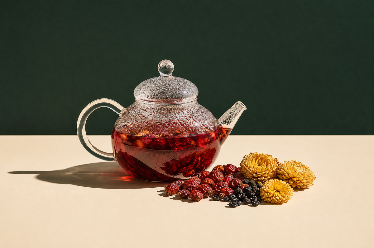 A round clear glass teapot of deep ruby-red rose hip and berry herbal tea beside a small pile of dried red rose hips