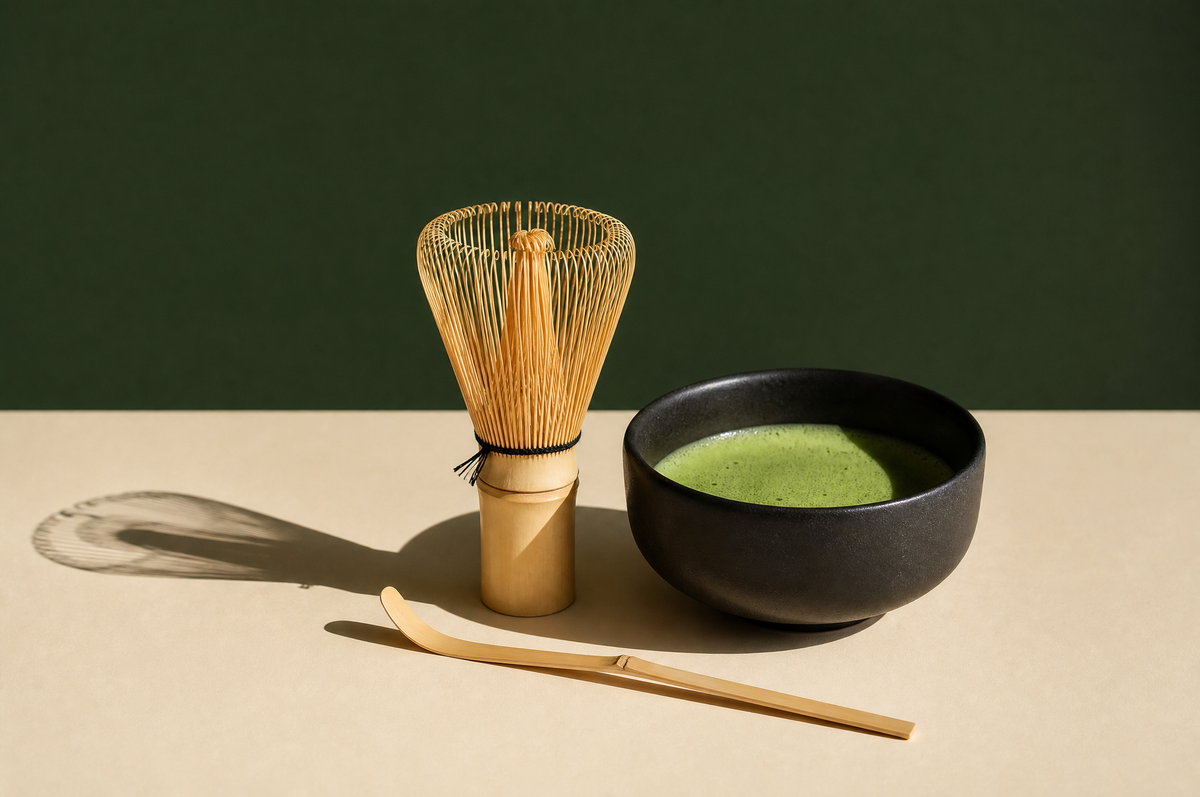 An upright bamboo matcha whisk with fine curled tines standing beside a curved bamboo tea scoop and a matte black ceramic