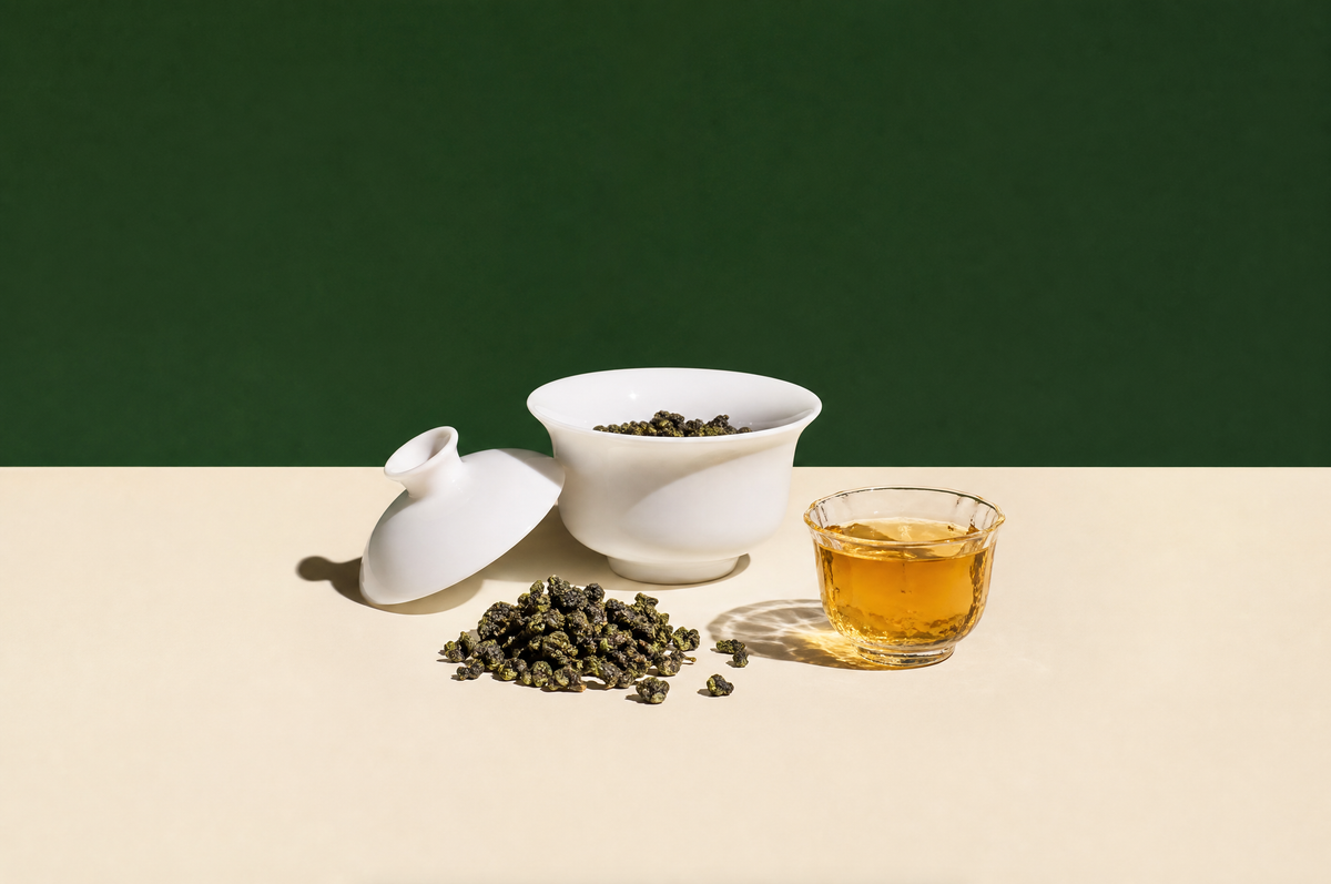 A white porcelain gaiwan with its lid resting ajar, a small heap of tightly rolled dark green oolong tea pearls