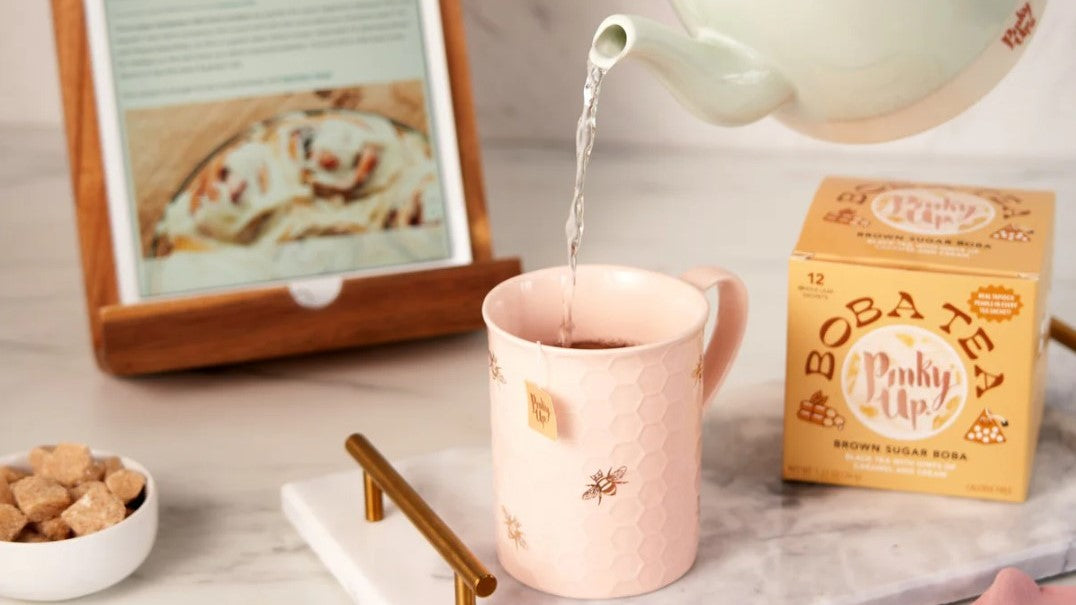 A light green teapot pours hot water into a pink, hexagon-patterned mug with bee motifs. 