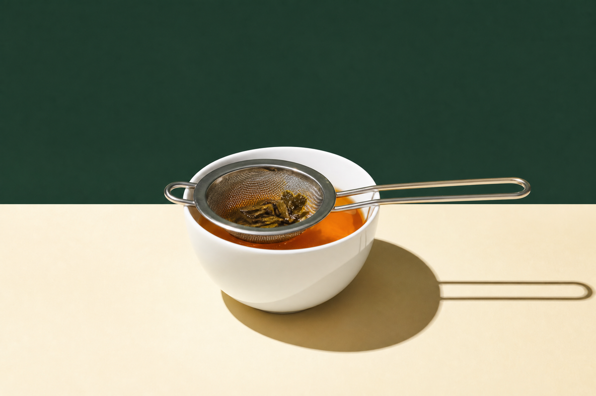 A fine-mesh stainless steel tea strainer with a long handle resting across the rim of a wide white porcelain cup of clear