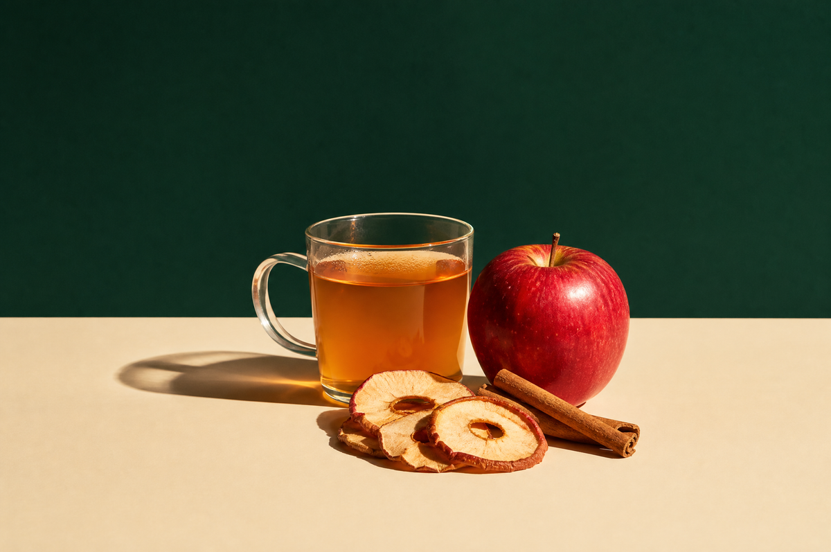 A clear glass cup of warm amber apple herbal tea beside one whole glossy red apple