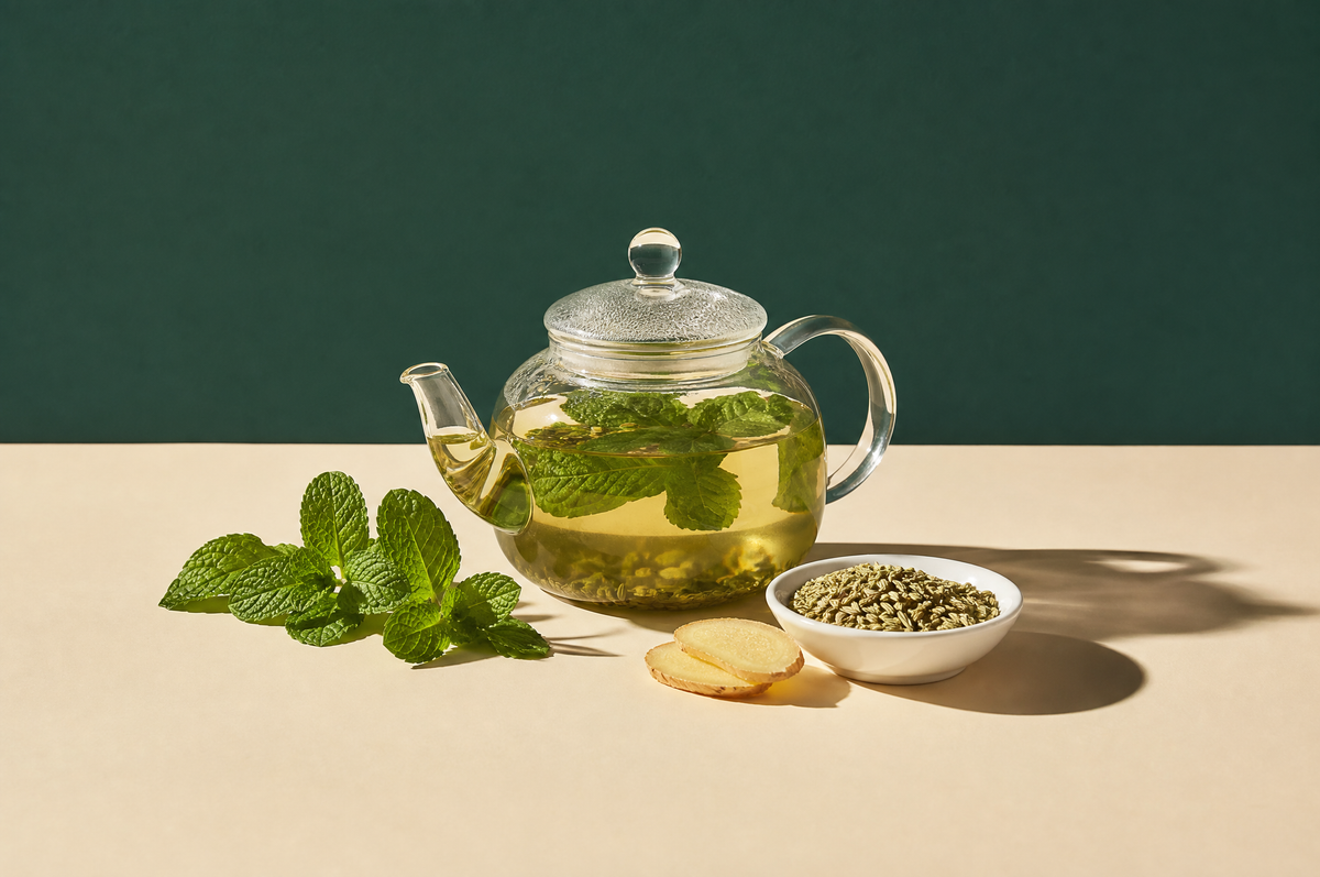A round clear glass teapot of pale green-gold peppermint and fennel herbal tea, next to a sprig of fresh peppermint leaves