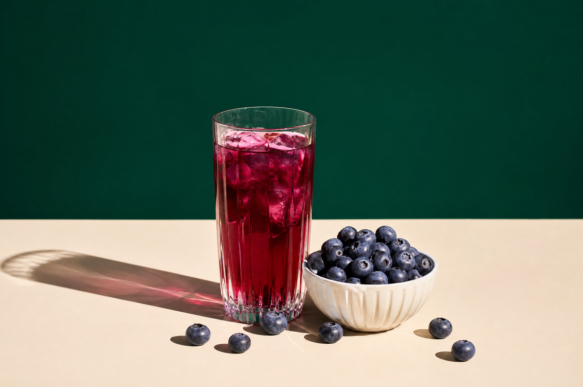 A tall clear glass tumbler of deep magenta-purple blueberry hibiscus tea beside a small white ceramic bowl heaped with fresh