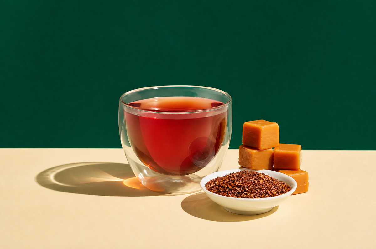 A clear glass double-walled cup of deep amber-red rooibos tea beside a small stack of glossy golden caramel cubes and a
