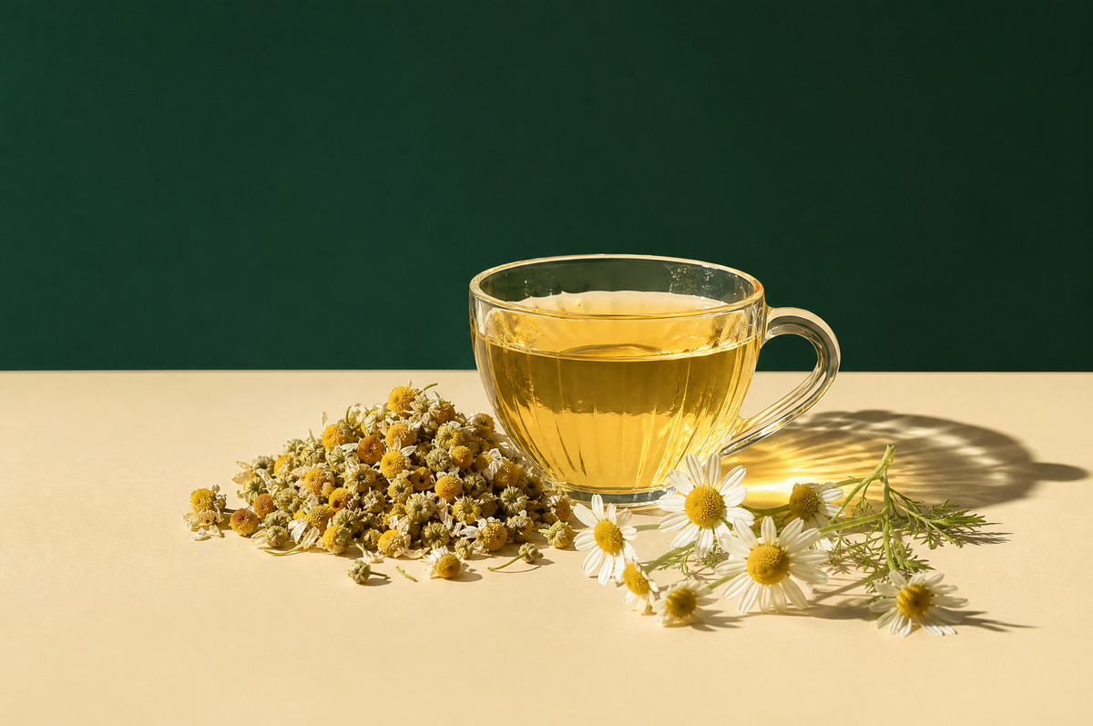 A clear glass teacup of pale golden chamomile tea beside a small heap of dried whole chamomile flowers with yellow centers