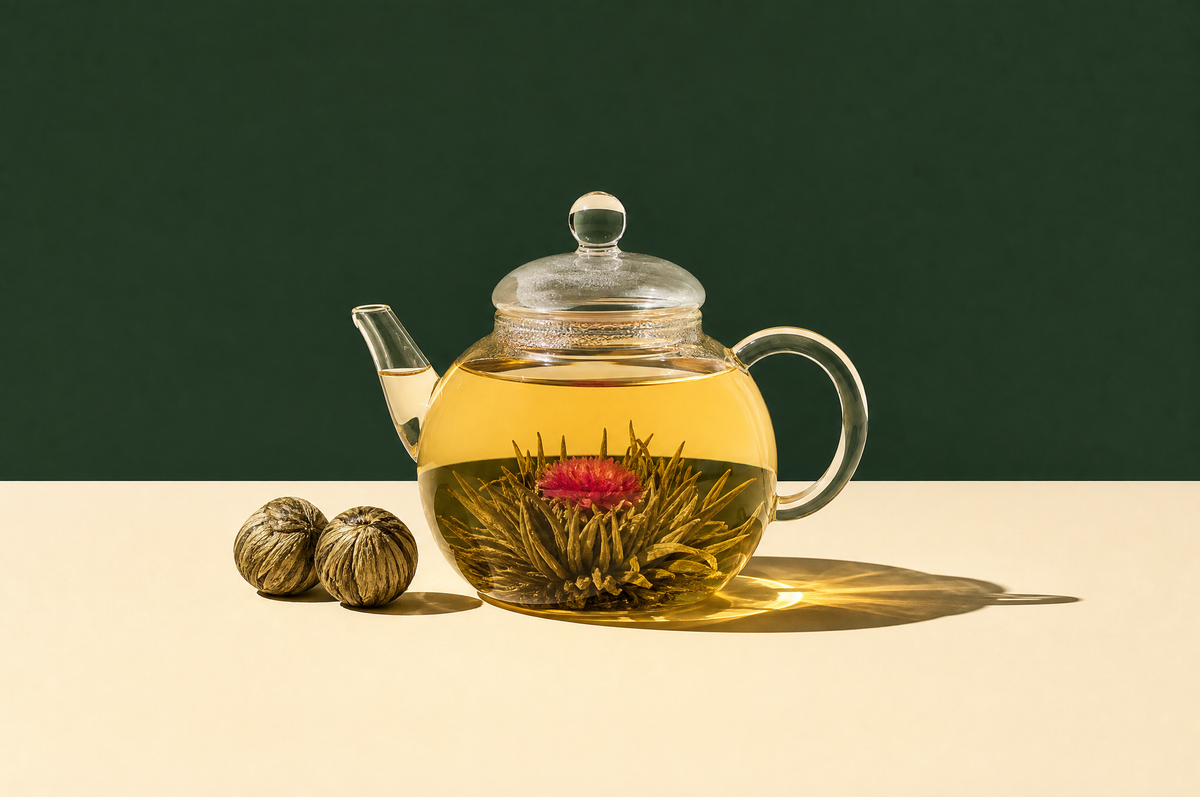 A round clear glass teapot filled with pale golden tea in which a blooming tea flower has opened into a green tea-leaf