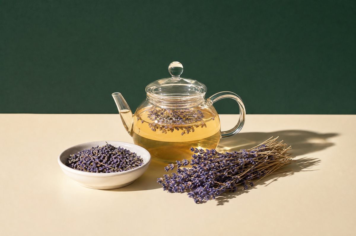 A small clear glass teapot of pale amber lavender tea next to a loose bundle of dried purple lavender sprigs and a small