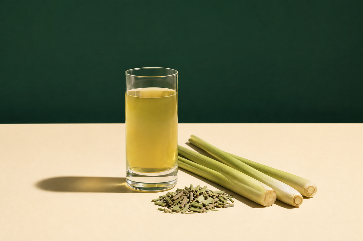 A tall clear straight-sided glass of pale greenish-yellow lemongrass tea beside three fresh green lemongrass stalks lying