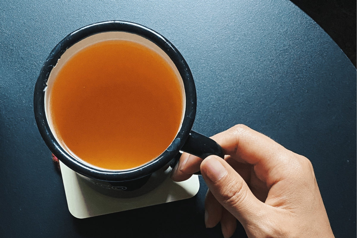 Hand holding a dark enamel mug of amber Passport tea on a dark table