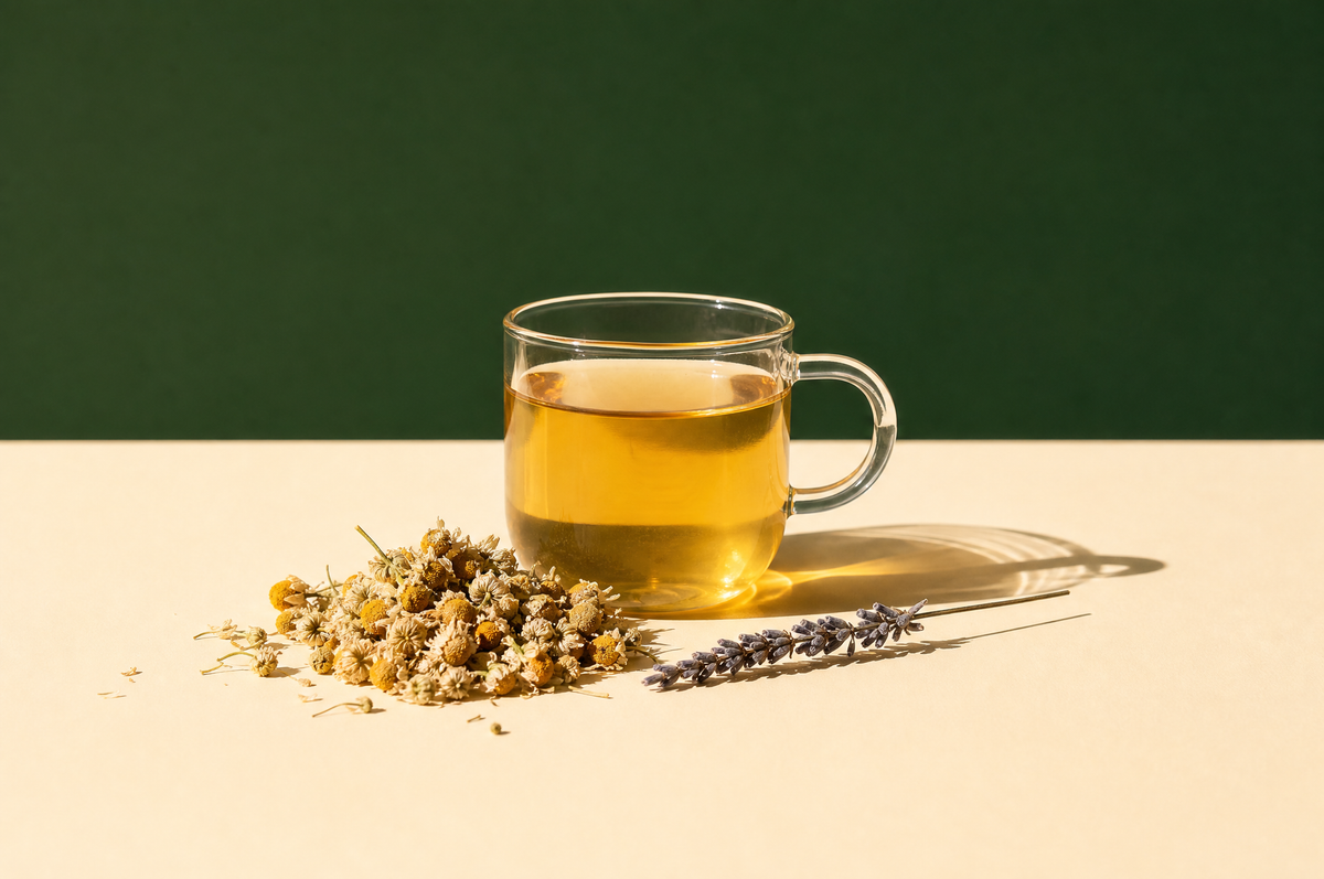 A clear glass mug of pale golden chamomile tea beside a small heap of dried whole chamomile flowers with yellow centers and