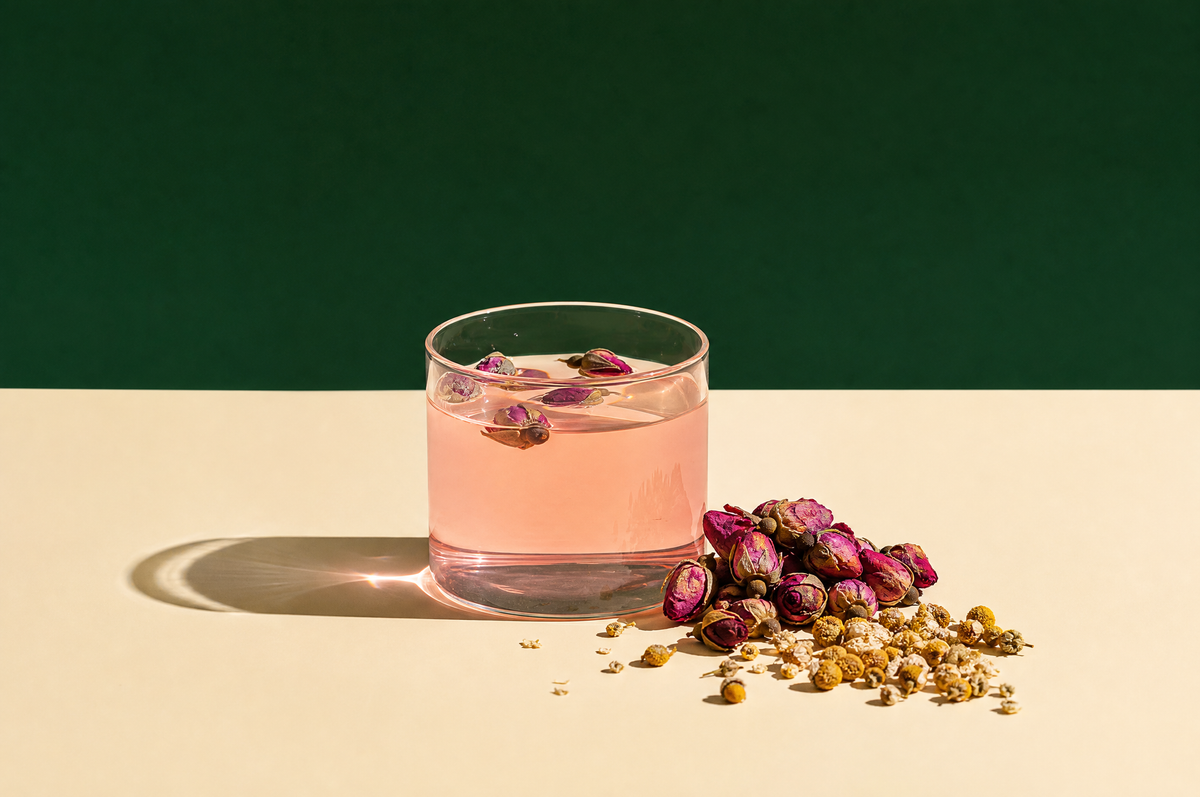 A clear handleless glass cup of pale rosy-pink rose herbal tea with a few dried rosebuds floating on top