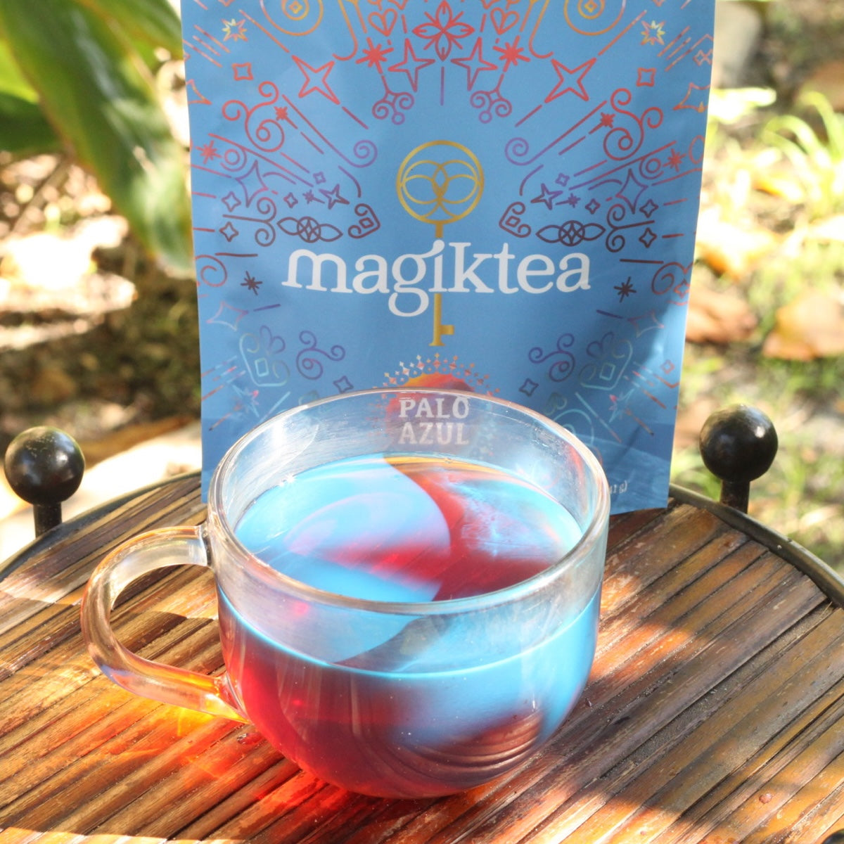 Glass mug with blue and red liquid next to a 'magiktea' package on a wooden surface.