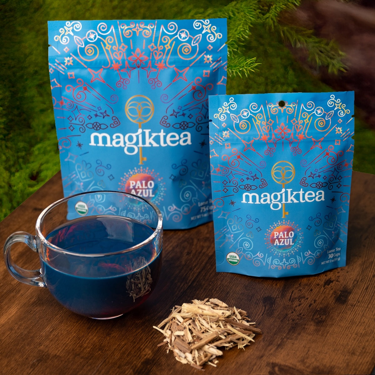 Two blue 'magiktea' packages with a glass of blue tea and dried herbs on a wooden surface.