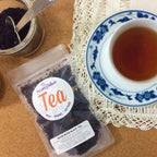 Plum Deluxe House Blend Black Tea (Creamy Vanilla English Breakfast) in a tea cup with a teapot in the background.