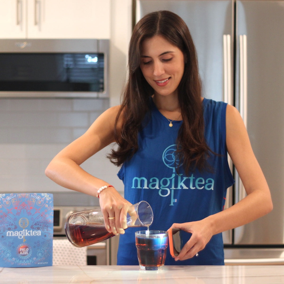Woman pouring tea from a bottle into a glass in a kitchen, wearing a blue 'magiktea' shirt.